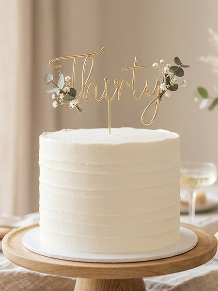 Gold wire "Thirty" cake topper with white floral accents displayed on a white frosted cake