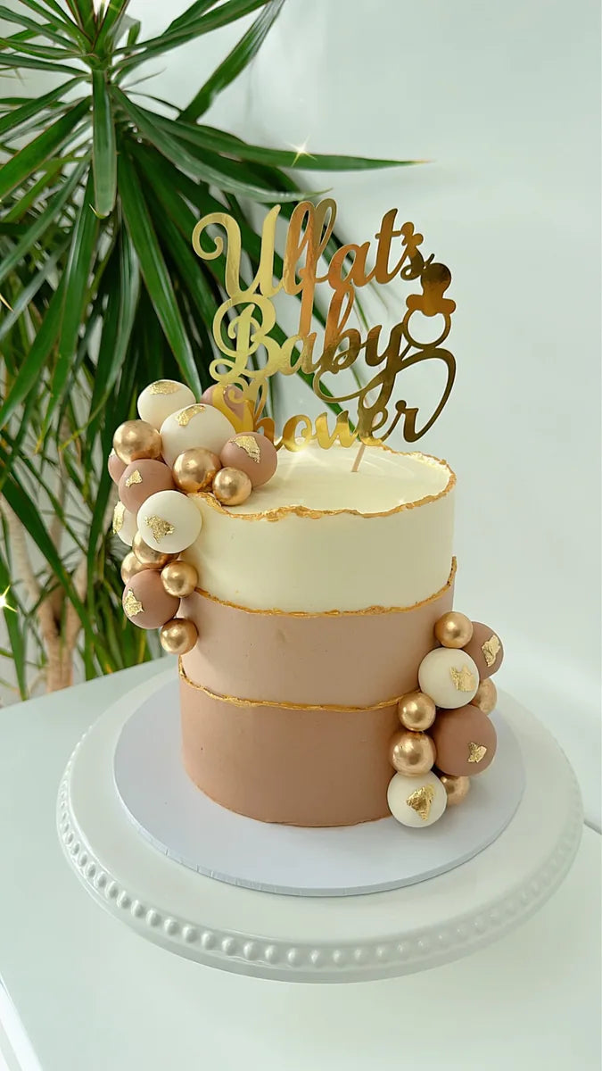 Gold personalised baby shower topper on a neutral cake with cream and gold decorations